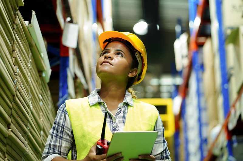 Female logistics worker with clipboard and safety apparel walks through the aisles of a warehouse.