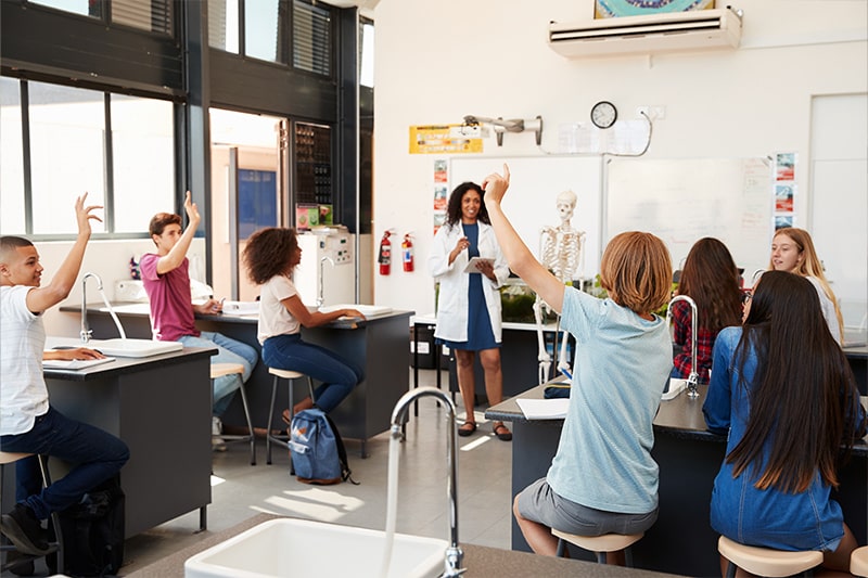 Science teacher lectures at the front of the classroom while students raise their hands.